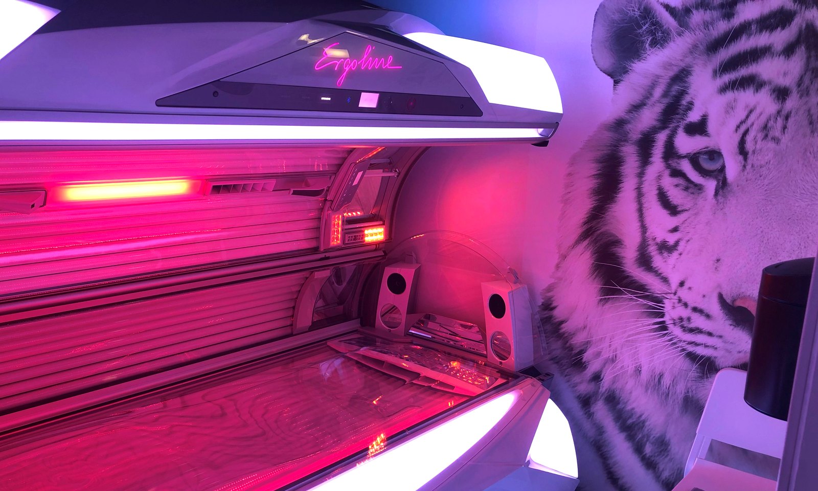 Een open Ergoline-zonnebank in Zonnestudio Leidschendam: roze gloed uit de lampen,
                  het Ergoline-neonlogo op de kap en een tijgerwand ernaast.